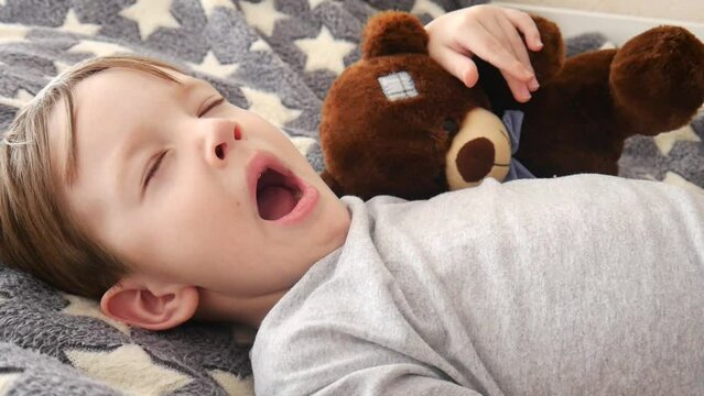 A pretty boy wakes up and yawns lying with teddy bear