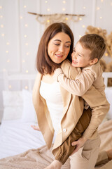 mom and baby boy play, cuddle and fool around at home on a bed in natural brown tones, a happy family with a small child has fun together
