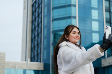 Urban portrait of young elegant business woman in winter casual clothes, white jacket. Walking in the city street, talking on the phone, making selfie. Working outdoor