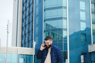 Urban portrait of young elegant business man in winter casual clothes, jacket, hat. Walking in the city street, talking on the phone, making selfie. Working outdoor