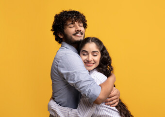 Portrait of affectionate indian couple hugging with closed eyes while standing isolated over yellow studio background
