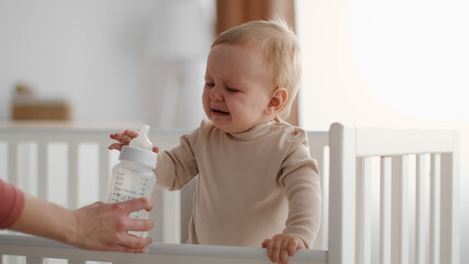 Cute little unhappy baby crying in crib, mother hand giving bottle with water or milk to toddler