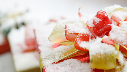 Closeup studio shot of Red paper wrapped present gift boxes with gold ribbon bowtie covered by white snow placed on wood table decorated with Christmas eve pine tree with sphere balls and snowflakes