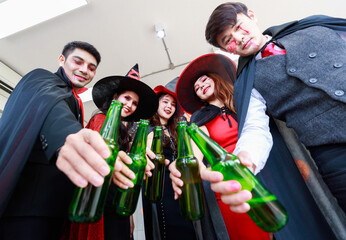 Portrait shot group of Asian young happy male and female friends in Halloween witch sorcerer ghost and skeleton costume with tall hat sitting on sofa holding beer bottles cheers together in party