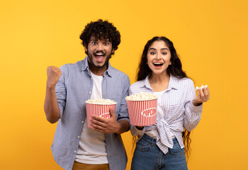 Excited indian man and woman eating popcorn, watching movie or football game, celebrating success...