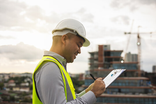 Inspector engineer man or architect with clipboard with white safety helmet in city construction site. Checking with checklist on rooftop building construction at capital.