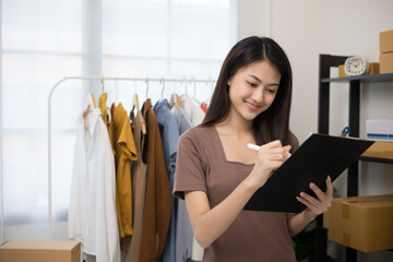 Young beautiful asian woman business owner at fashion store holding clipboard check stock on rails clothing. Asian female entrepreneur working in clothes shop.