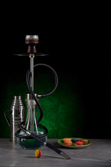 Obraz na płótnie Canvas Beautiful modern hookah standing on a table surrounded by a metal protective cap and a plate with colorful macaroons on a dark green background. Space for copying. Vertical location.