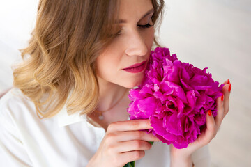 Fototapeta premium woman in white shirt, holding peony bud in her hands, closed her eyes. Portrait of beautiful blonde woman. Surprise bouquet of flowers for date, mothers day or valentines day.
