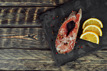 Raw salmon steak on a black stone plate. Red salmon with lemon close up. Food on shabby wooden table.