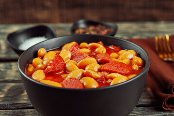 Beans baked with sausages. Beans with fried sausages in tomato sauce. Food in a black bowl on a brown wooden table.
