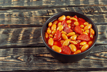 Beans baked with sausages. Beans with fried sausages in tomato sauce. Food in a black bowl on a brown wooden table. Copy space and free space for text near food.