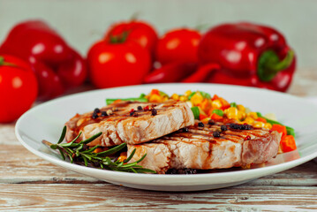 Pieces of meat grilled. Steak with rosemary and pepper in a white plate. Meat dish with red sauce on a white wooden table. Salad of corn, carrots and green peas near the steak.
