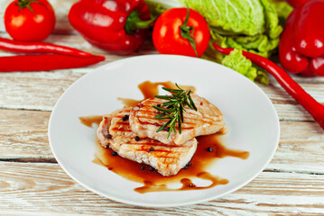Pieces of meat grilled. Steak with rosemary and pepper in a white plate. Meat dish with red sauce on a white wooden table. Fresh tomatoes, peppers and cabbage near the plate.