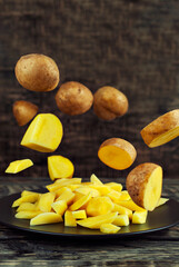 Raw potatoes fall on a black plate. Potatoes fly over a wooden table. Whole and sliced potatoes. Healthy food and farming concept.