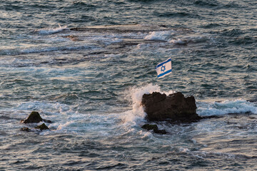 A large, blue and white flag of the State of Israel perched on a rock in the Mediterranean Sea off...