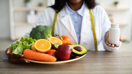 Dietitian offering choice between fresh fruits and vegetables and tablets at clinic, holding jar of...