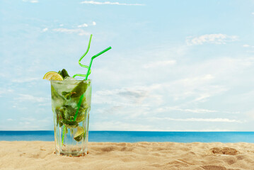 Green cocktail with a straw on the sand. Alcoholic mojito with ice, lime and mint on the beach. Sunset, sea and sky in the background. Copy space and free space for text near the glass.