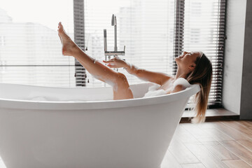 Playful young lady shaving legs with razor, lying in foamy bubble bath, enjoying domestic skin care...
