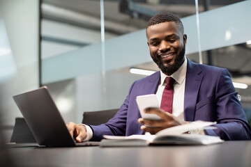 Cheerful black manager using smartphone while working on laptop