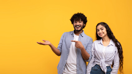 Cheerful indian couple pointing at open palm, man demonstrating invisible object, showing copy space, panorama