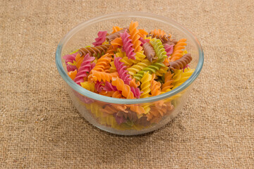 Uncooked colored spiral pasta in glass bowl on burlap surface