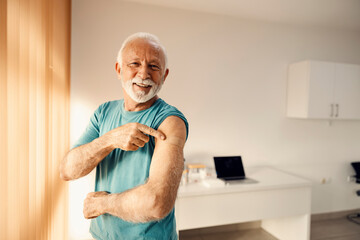 A happy senior man in doctor's office pointing at his vaccinated shoulder. Immunization and vaccination. - obrazy, fototapety, plakaty