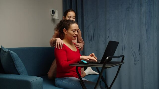 Multiethnic woman working at home, using laptop while lovely girl cuddling busy mother, asking to make a break and give attention to child at home. Home office and motherhood at same time.
