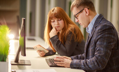 Coworking, a young woman and a business man are discussing a project looking at a laptop monitor and screen, studying documentation, discussing project activities, business partners.