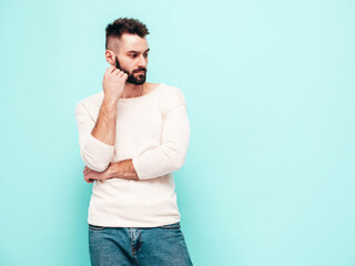 Portrait of handsome confident stylish hipster lambersexual model.Man dressed in white sweater and jeans. Fashion male isolated on blue wall in studio