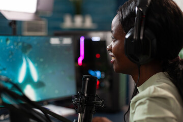 Concentrated cyber gamer woman talking into microphone during live streaming with remote player discussing online games championship. Player playing space shooter videogames using RGB computer