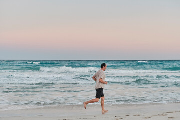 Senior asian man running barefoot on sandy ocean beach at sunrise.