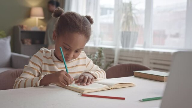 Waist up slowmo of 5 year old African-American girl drawing in copybook sitting at desk in cozy living room spending leisure time with her mom