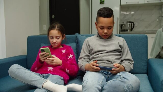 Diverse multiethnic kids using smartphones sitting on sofa indoors. Moving shot. Multinational children holding digital devices, having fun with mobile app, playing game, checking social media at home