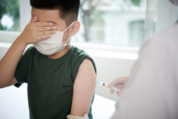 Little boy in mask closing his eyes with hands while doctor giving an injection from covid at...
