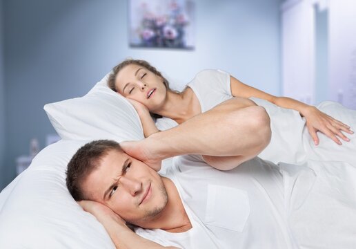 Despaired millennial man on bed covers ears and suffers from snoring of sleeping wife