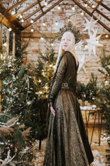 young albino woman with blue eyes and long white hair in beautiful green dress and crown stands in loft room decorated with wooden greenhouse and Christmas trees with twinkle lights, diverse people