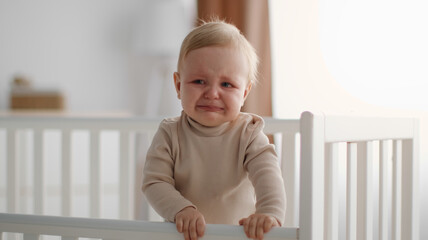 Baby unhappiness. Portrait of adorable unhappy crying baby standing in crib, suffering from colics...
