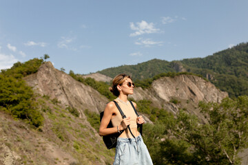 Naklejka premium Adorable charming lady with wavy light hair wearing t-shirt and shorts with backpack is moving against green mountains in sunlight in warm spring day. Woman traveling in Woman's Day