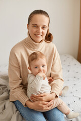 Portrait of smiling positive woman with ponytail wearing beige sweater with infant baby, young mother sitting on bed and looking at camera with happy facial expression.