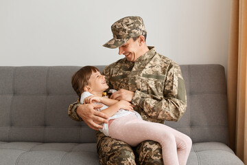 Horizontal shot of positive happy soldier man in camouflage uniform sitting with daughter on sofa,...