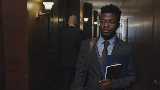 Waist up shot of African American businessman greeting colleague while walking with file folder and notepad through corridor of office center
