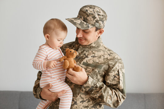 Portrait of soldier man wearing camouflage holding his infant daughter and giving her little teddy bear toy, present after returning home from army or war.