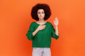 I promise! Serious woman with Afro hairstyle wearing green sweater standing raising hand and saying swear, making loyalty oath, pledging allegiance. Indoor studio shot isolated on orange background.