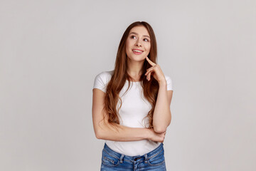 Portrait of adorable dreamy woman standing looking away, keeping finger on cheek, dreaming, making wish, love, wearing white T-shirt. Indoor studio shot isolated on gray background.