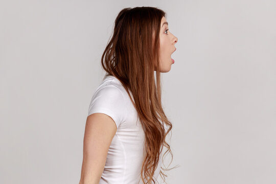 Side view portrait of astonished amazed woman looking with mouth open in surprise, shocked facial expression, wearing white T-shirt. Indoor studio shot isolated on gray background.