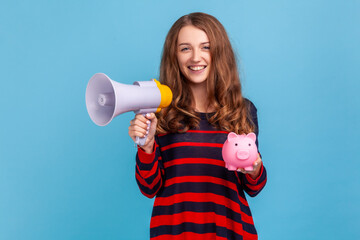 Portrait of positive woman wearing striped casual style sweater, holding piggy bank and megaphone, looking at camera with toothy smile. Indoor studio shot isolated on blue background.