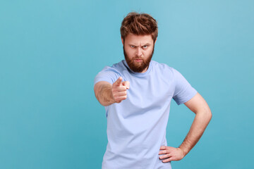 Hey you. Portrait of young adult displeased bossy bearded man pointing finger to camera and blaming for mistakes, shaming making you guilty. Indoor studio shot isolated on blue background.