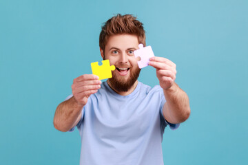 Portrait of smiling bearded man holding two puzzle parts and smiling joyfully, ready to connect jigsaw pieces, symbol of union and association. Indoor studio shot isolated on blue background.