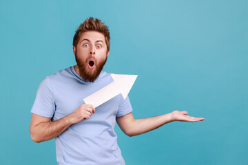 Portrait of bearded man holding arrow pointing up looking at camera with open mouth, growth and increase concept, presenting copy space. Indoor studio shot isolated on blue background.